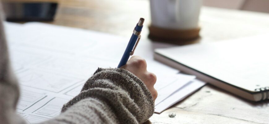 person writing on brown wooden table near white ceramic mug